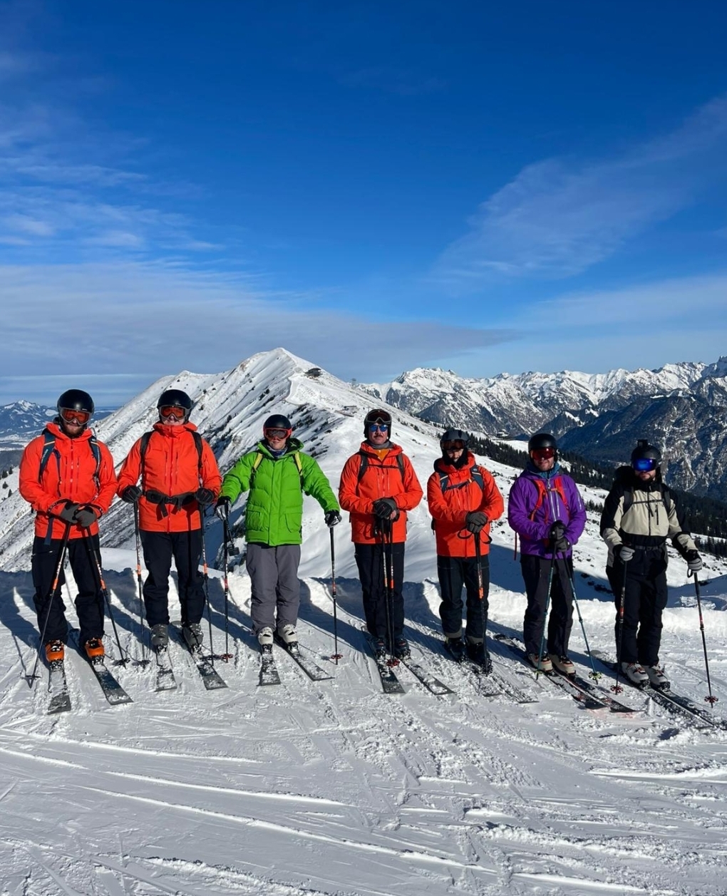 Group of skiers lined up on a snowy ridge in the Bavarian Alps under a clear blue sky.