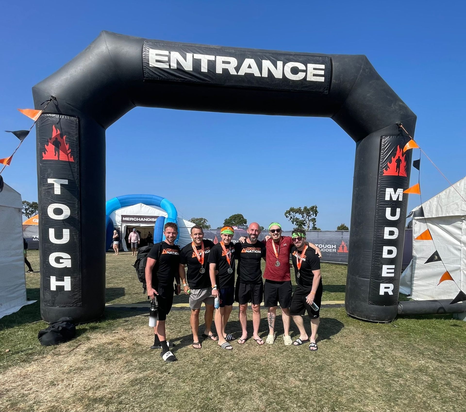 Team of six posing under the Tough Mudder entrance arch with finisher medals.