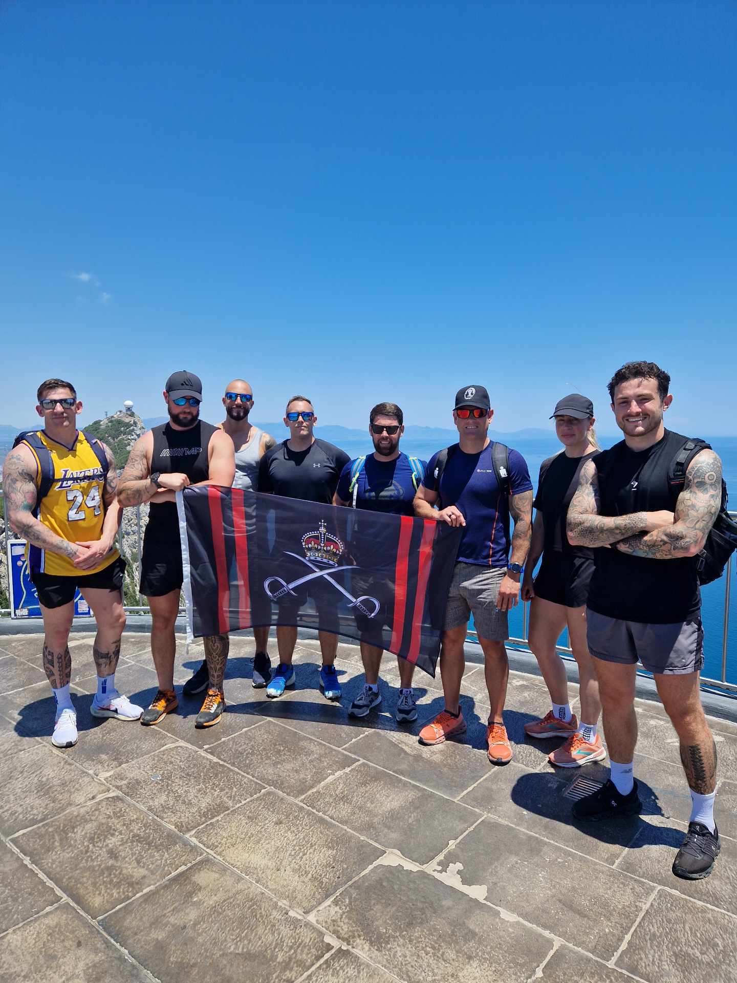 Group of eight on the upper Rock of Gibraltar holding a regimental flag with the Mediterranean behind them.